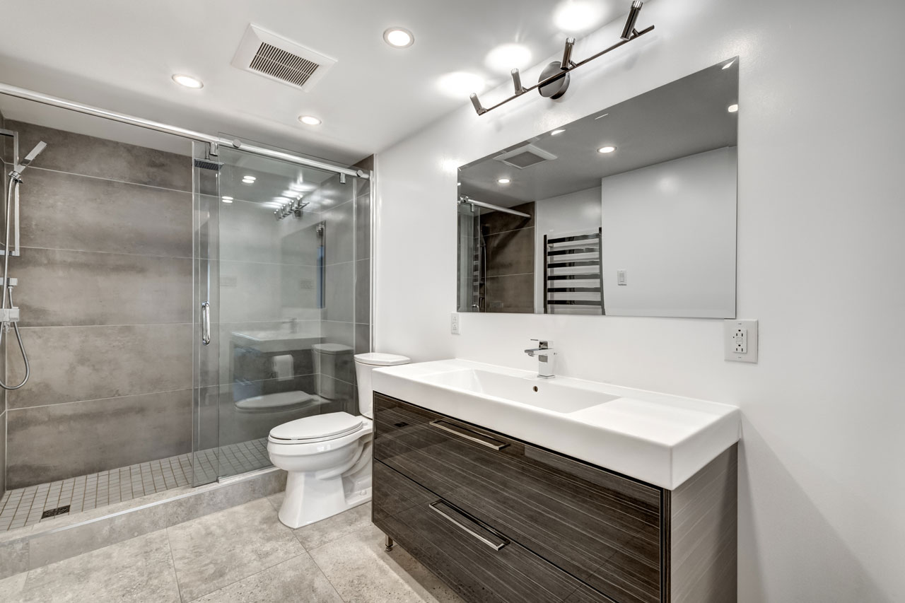 Stylish bathroom featuring a large, frameless glass-enclosed shower with dark grey tiles. The space includes a modern vanity with a long white sink and dark wood cabinets, complemented by a large mirror and minimalist lighting. The overall design is clean and contemporary, emphasizing functionality and modern aesthetics.