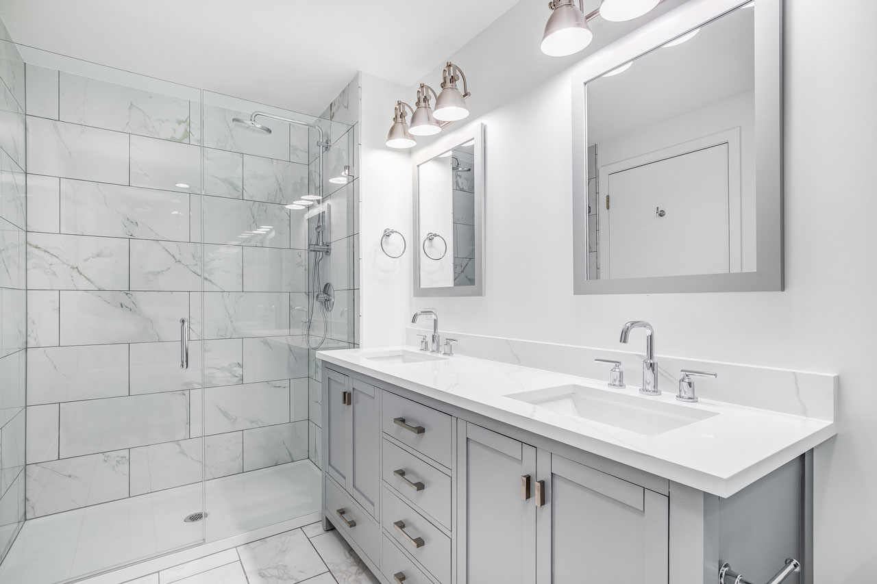 Modern bathroom featuring a double vanity with sleek white countertops and grey cabinets, complemented by matching framed mirrors. The space includes a glass-enclosed shower with white marble tiles, creating a clean and elegant look. Overhead lighting and chrome fixtures add to the bright and polished ambiance.