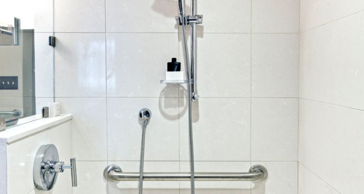 Brightly lit bathroom corner showcasing a practical shower setup with large white tiles. It features a stainless steel adjustable shower head, a hand-held shower option, and multiple safety grab bars, ensuring accessibility and security. A niche holds toiletries within easy reach, reflecting a thoughtful design for user convenience and safety.