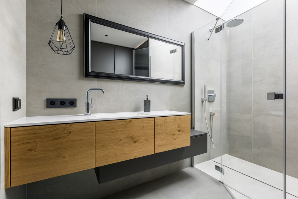 Contemporary bathroom featuring a floating vanity with natural wood drawers and a white countertop. The room includes a glass-enclosed shower with large grey tiles and minimalist fixtures. A black framed mirror, geometric pendant light, and clean lines contribute to the modern industrial aesthetic, creating a stylish and functional space.