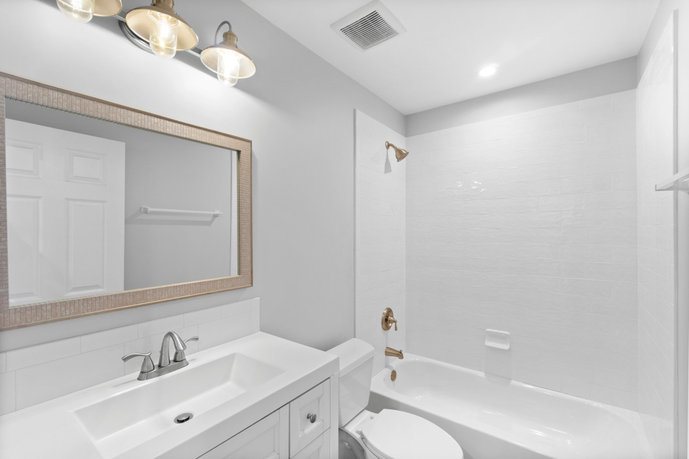 Bright bathroom featuring a classic white vanity with a rectangular sink, complemented by a large framed mirror and overhead lighting. The space includes a bathtub with white tiled walls and modern brushed nickel fixtures. The minimalist design and soft lighting create a clean and inviting atmosphere.