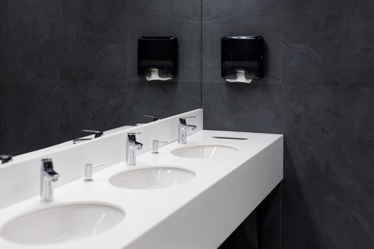 Contemporary bathroom featuring a long white countertop with multiple sinks, set against a dramatic dark gray slate tile wall. Chrome faucets add a sleek, modern touch to the minimalistic design. Above the sinks, automated paper towel dispensers are mounted on the wall, emphasizing functionality and cleanliness in this stylish space.